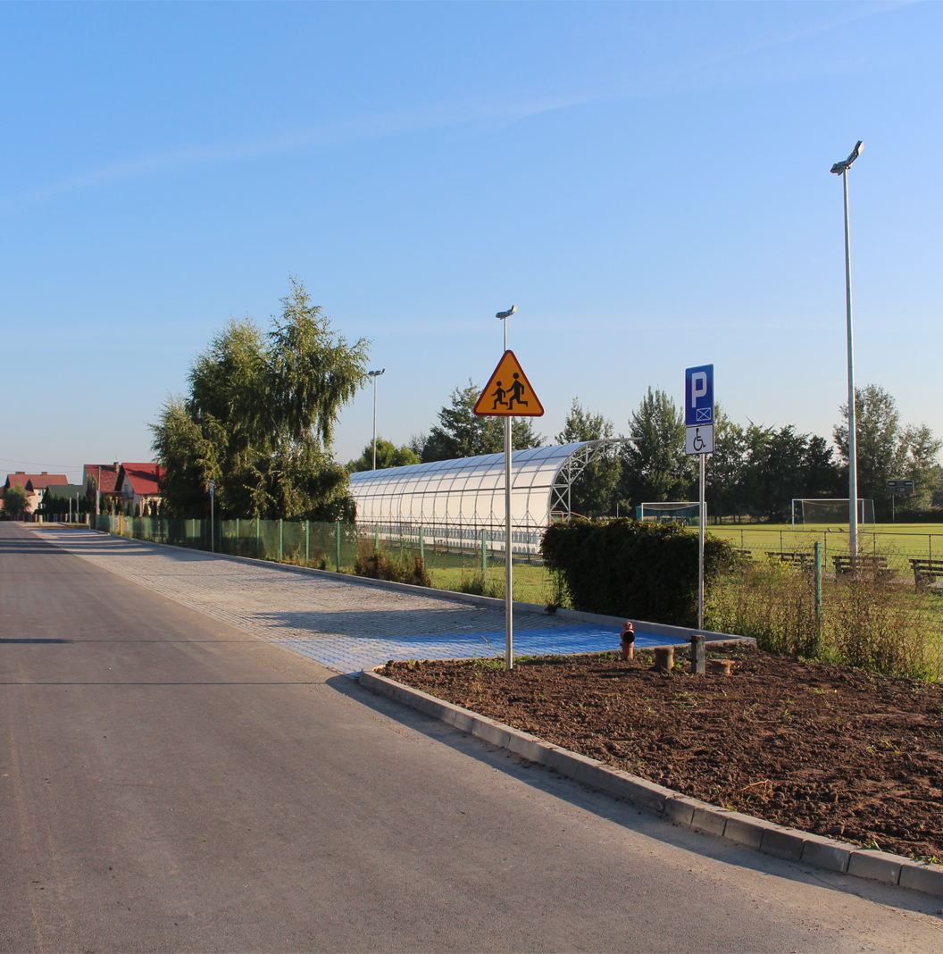 Sokolniki. Droga asfaltowa przy stadionie. w tle miejsce miejsce parkingowe dla osoby niepełnosprawnej, dalej widać trybunę sportową